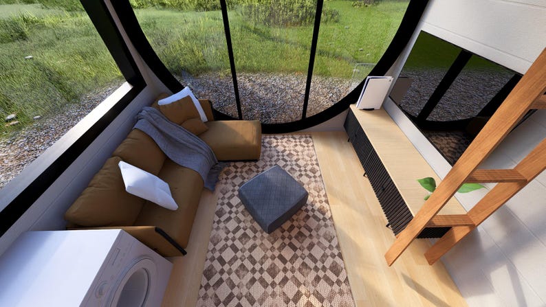 May include: Interior view of a modern living space with large windows overlooking a green landscape. The room features a brown sectional sofa with white pillows, a patterned rug, and a wooden ladder. A small gray ottoman sits on the rug.