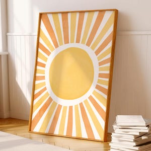 May include: A framed art print with a sun design. The sun has a yellow centre and orange and white rays. The artwork is in a wooden frame and leans against a white wall. The room has a light-coloured rug and hardwood floors.