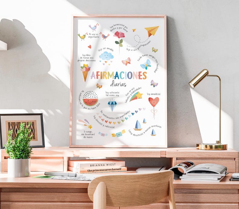 May include: A framed print with the words "AFIRMACIONES diarias" in colorful lettering. The print features various illustrations, including birds, butterflies, and a watermelon slice. The print is displayed on a wooden desk with a small plant and books.