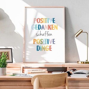 Peut inclure: Une impression encadr&eacute;e avec le texte "POSITIVE GEDANKEN schaffen POSITIVE DINGE" en lettres color&eacute;es sur fond blanc. L'impression est accroch&eacute;e &agrave; un mur blanc au-dessus d'un bureau en bois.