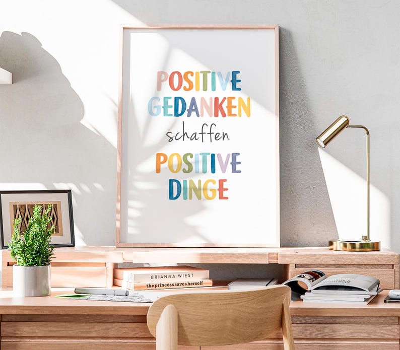 Peut inclure: Une impression encadr&eacute;e avec le texte "POSITIVE GEDANKEN schaffen POSITIVE DINGE" en lettres color&eacute;es sur fond blanc. L'impression est accroch&eacute;e &agrave; un mur blanc au-dessus d'un bureau en bois.