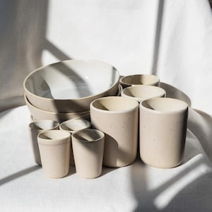 Könnte beinhalten: Eine Sammlung beigefarbener Keramikgeschirr, darunter Schalen und verschieden große Tassen. Die Schalen sind gestapelt und die Tassen in einer Gruppe angeordnet. Die Keramikteile haben eine gesprenkelte Textur und ein mattes Finish.