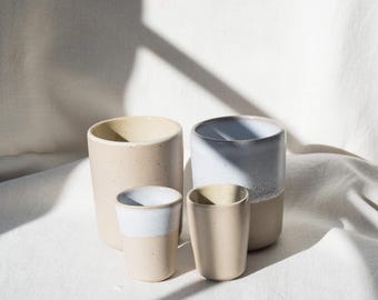May include: Four ceramic cups in different sizes and shapes. The cups are mainly beige with white or light blue rims. The cups are arranged on a white surface, with shadows adding depth and texture.