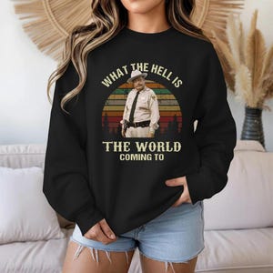 May include: Black sweatshirt featuring a vintage-style graphic of a man in a sheriff's uniform against a sunset backdrop. The text reads "WHAT THE HELL IS THE WORLD COMING TO".