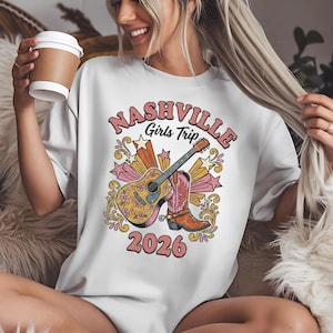 May include: White t-shirt with a colorful graphic design featuring a guitar, cowboy boots, and the text "Nashville Girls Trip 2026". The design includes stars and floral elements, creating a vintage aesthetic.