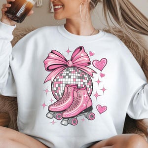 May include: White sweatshirt with a pink disco ball, bow, hearts, and roller skates. The design includes pink roller skates with black wheels, a large pink bow, and several pink hearts. The sweatshirt is a casual, comfortable style.