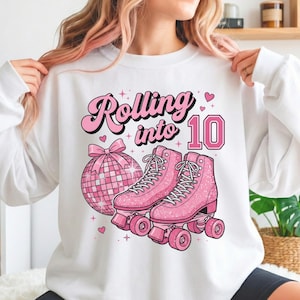 May include: White sweatshirt featuring a pink graphic with the text "Rolling into 10". The design includes a disco ball, pink roller skates, and hearts. A casual, comfortable style.
