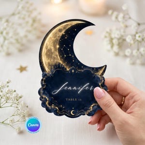 May include: A crescent moon-shaped place card featuring a dark blue background with gold celestial patterns, stars, and constellations. The card displays the name Jennifer in white script and the text TABLE 13 in a simple gold font.