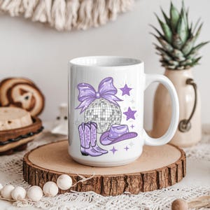May include: White ceramic mug with a purple glitter design featuring a disco ball, cowboy hat, and boots. The design also includes a purple bow and stars.