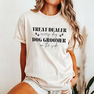 以下が含まれることがあります： クリーム色のTシャツに、黒い文字で「Treat Dealer every day Dog Groomer on the side」と書いてあります。テキストは肉球のプリントで囲まれています。