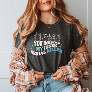 Puede incluir: Una camiseta gris oscuro con un gráfico de esqueletos bailando y el texto "You inspire my inner serial killer" en colores pastel.