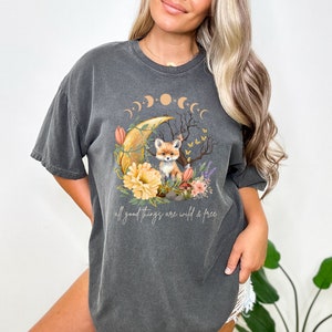 May include: A charcoal gray t-shirt with a graphic of a fox sitting on a crescent moon with flowers and butterflies. The text "all good things are wild & free" is printed below the graphic.