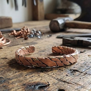 May include: A handcrafted copper cuff bracelet with a woven design, featuring alternating copper and brass segments. The open-ended bracelet sits on a wooden workbench, with tools and metal scraps in the background.