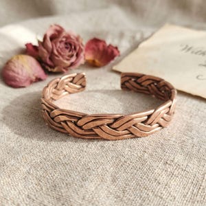 May include: A copper-colored cuff bracelet with a braided design. The open-ended bracelet is made of interwoven metal strips. Dried rose petals and a vintage paper add to the aesthetic. The bracelet is a piece of jewelry.