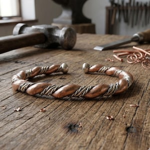 May include: A copper and silver cuff bracelet with a twisted design. The bracelet has a copper base with silver wire wrapped around it. The ends of the bracelet have silver ball accents. The bracelet is on a wooden surface.