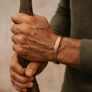 May include: A copper-coloured cuff bracelet with a diagonal grooved design. The bracelet is worn on a wrist, and the person is holding a wooden stick. The bracelet is a simple, yet stylish accessory.