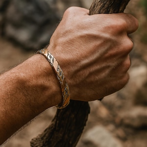 May include: A metallic cuff bracelet featuring a polished, braided texture with alternating smooth and intricate woven sections. The bracelet has a warm, golden-bronze tone and a rigid, open-ended design that wraps around the wrist.