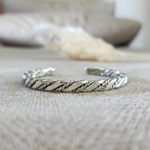 Könnte beinhalten: Ein silberfarbenes Armband in Manschettenform mit einem gedrehten Seildesign. Das Metallband hat eine polierte Oberfläche und eine offene Struktur, die ein sich wiederholendes Muster aus ineinander verschlungenen Strängen zeigt.