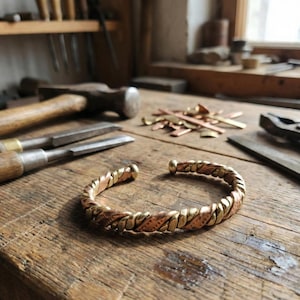 Può includere: Un bracciale a fascia realizzato con fili intrecciati di rame e ottone. Il bracciale presenta un design intrecciato e testurizzato con tonalità metalliche alternate di rame bruno-rossastro e ottone giallo brillante, formando un cerchio aperto.