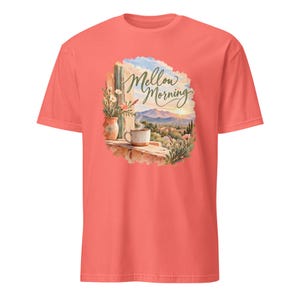 Op de afbeelding: Een koraalkleurig t-shirt met korte mouwen en een grafische print van een landschap met een dampende mok en een vaas met bloemen op een richel. De tekst Mellow Morning staat in cursief schrift boven de bergen en de zonsopgang.