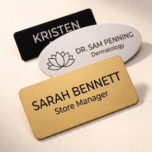 May include: Three rectangular and oval name badges. One is black with white text reading KRISTEN. One is silver oval with a lotus icon and text DR. SAM PENNING Dermatology. One is gold with black text reading SARAH BENNETT Store Manager.