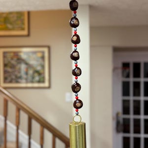 May include: A hanging wind chime featuring a string of dark brown, round objects interspersed with red and gray beads. A gold-colored metal bell hangs at the bottom. The chime is suspended from a white ring.