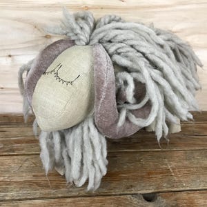 May include: A plush sheep toy with a light gray woolly mane and a beige face. The sheep has long, floppy ears in a muted purple color and embroidered closed eyes. The toy is resting on a wooden surface.