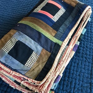May include: Folded patchwork quilt with a variety of fabrics in shades of blue, brown, green, and cream. The quilt features a log cabin design with a mix of solid and patterned fabrics, including houndstooth. The edges are finished with a red and cream fabric.