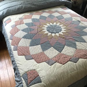May include: A handmade quilt with a floral design in shades of pink, blue, cream, and brown. The quilt features a central starburst pattern and is placed on a bed with a matching blue comforter and pillow.