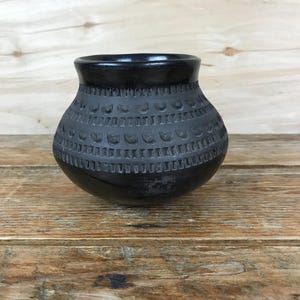May include: A small, black ceramic vase with a rounded body and a slightly flared rim. The vase features a textured pattern of repeating shapes around its midsection. The vase sits on a wooden surface.