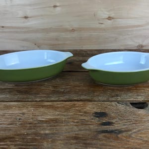 May include: Two oval-shaped, green ceramic baking dishes with white interiors. Each dish has a handle on either side. The dishes are set on a wooden surface, with a light-colored wooden background.
