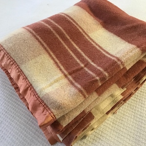 May include: Folded wool blanket with alternating stripes of cream and muted red-brown. The blanket has a satin edge in a matching red-brown color. The blanket is folded neatly, showing the texture and pattern.