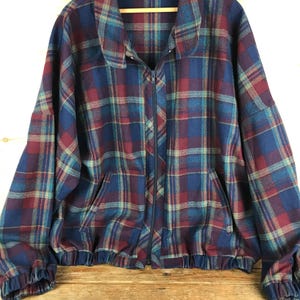 May include: A vintage plaid jacket in shades of blue, burgundy, and teal. The jacket features a full zipper, two front pockets, and elastic cuffs and hem. The jacket is hanging on a wooden hanger.
