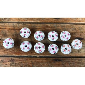 May include: Ten round white ceramic knobs, each featuring a central hole and a decorative pattern of pink lotus-like flowers accented with black swirling lines and small black dots arranged in a circular design around the center opening.