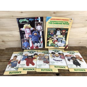 May include: A collection of Cabbage Patch Kids sewing pattern envelopes and booklets featuring illustrations of dolls in various outfits. The paper covers display colorful images of dolls wearing knitted sweaters, hoodies, dresses, and overalls with the Butterick brand logo.