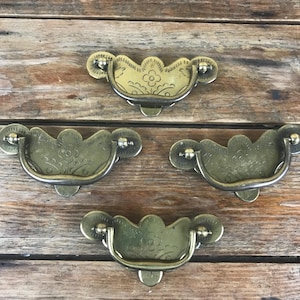 May include: Four antique brass drawer pulls with a floral design. Each pull has a curved handle and decorative backplates. The pulls are arranged on a weathered wooden surface, showcasing their vintage aesthetic.