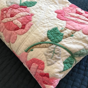 May include: A white quilted throw blanket featuring large pink rose appliques with green and grey leaves. The fabric has a textured, stitched pattern throughout, showcasing detailed floral motifs with layered petals and embroidered stems.