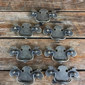 May include: A set of six antique-style brass drawer pulls featuring ornate, embossed floral patterns on the backplates and curved, swinging handles. Each piece has a weathered, dark metallic finish with intricate detailing and symmetrical shapes.