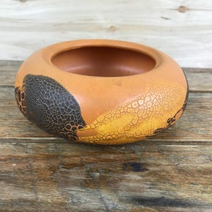 May include: A round, terracotta-colored bowl with a wide rim. The exterior features a textured pattern in shades of black, yellow, and orange. The bowl is sitting on a wooden surface.