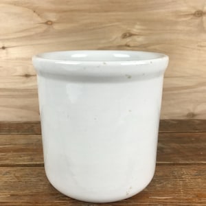 May include: A white ceramic crock with a rounded base and a slightly flared rim. The cylindrical container has a smooth, glossy finish and appears to be empty. The background is a wooden surface.