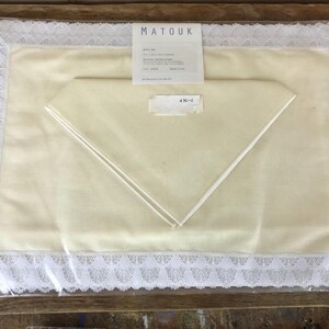 May include: A white tablecloth with a lace border and a white napkin folded into a triangle. The tablecloth is packaged in plastic and has a label that says "Matouk" and "4 N/N1".