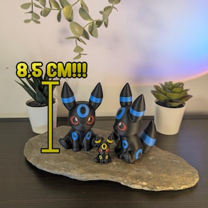 Può includere: Tre statuette nere con dettagli blu e rossi, tra cui due figure di Umbreon e una piccola figura di Eevee, sono esposte su una pietra. La figura più alta misura 8,5 cm di altezza. Le figure sono poste su uno sfondo di piccole piante in vaso.