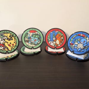 May include: Four circular coasters with Pokémon characters. Each coaster has a unique design featuring Pikachu, Bulbasaur, Charmander, and Squirtle. The coasters are on small stands. The background is a dark brown surface.