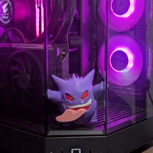 Op de afbeelding: Een paars Gengar-beeldje met een roze tong in een computerkast. De kast heeft een zwart frame en heldere glazen panelen. De computer heeft paarse LED-verlichting en ventilatoren.