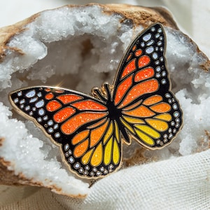 May include: A butterfly-shaped enamel pin featuring a monarch butterfly design with vibrant orange, yellow, and black sections. The wings are filled with a sparkling glitter finish and accented by small white dots along the black outer edges.