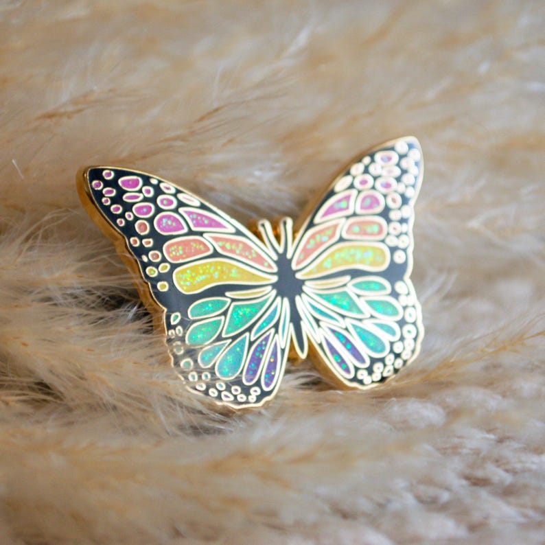 May include: A gold-toned butterfly pin with iridescent wings. The wings feature a black outline with colorful sections of pink, yellow, and teal. The pin is set against a soft, neutral background.