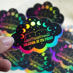 May include: Holographic cloud-shaped sticker with a rainbow gradient edge. The sticker features a starry night sky design with moons, stars, and an open book. Text reads "Escapism at its Finest".