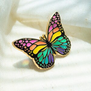 May include: A colorful butterfly pin with a gold-tone outline. The butterfly's wings feature a vibrant array of colors, including pink, orange, yellow, green, and purple, set against a black background. The pin is a decorative accessory.