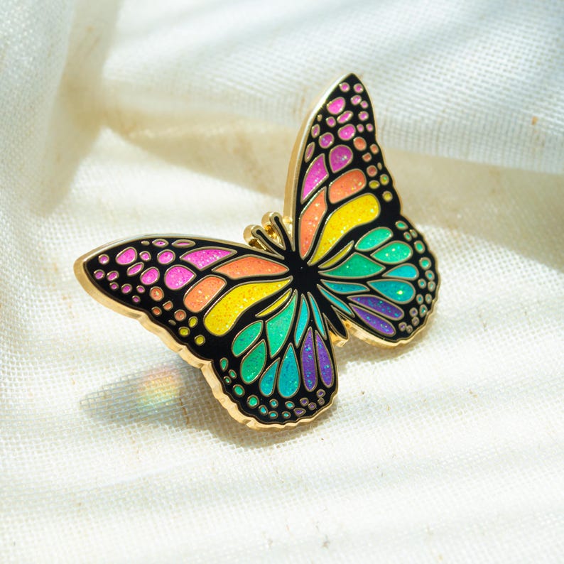 May include: A colorful butterfly pin with a gold-tone outline. The butterfly's wings feature a vibrant array of colors, including pink, orange, yellow, green, and purple, set against a black background. The pin is a decorative accessory.