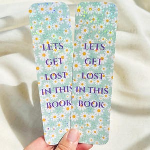 May include: Two daisy-patterned bookmarks with the text "LETS GET LOST IN THIS BOOK" in purple. The bookmarks have a light green background and a glittery finish. The daisy pattern includes white petals and yellow centers.